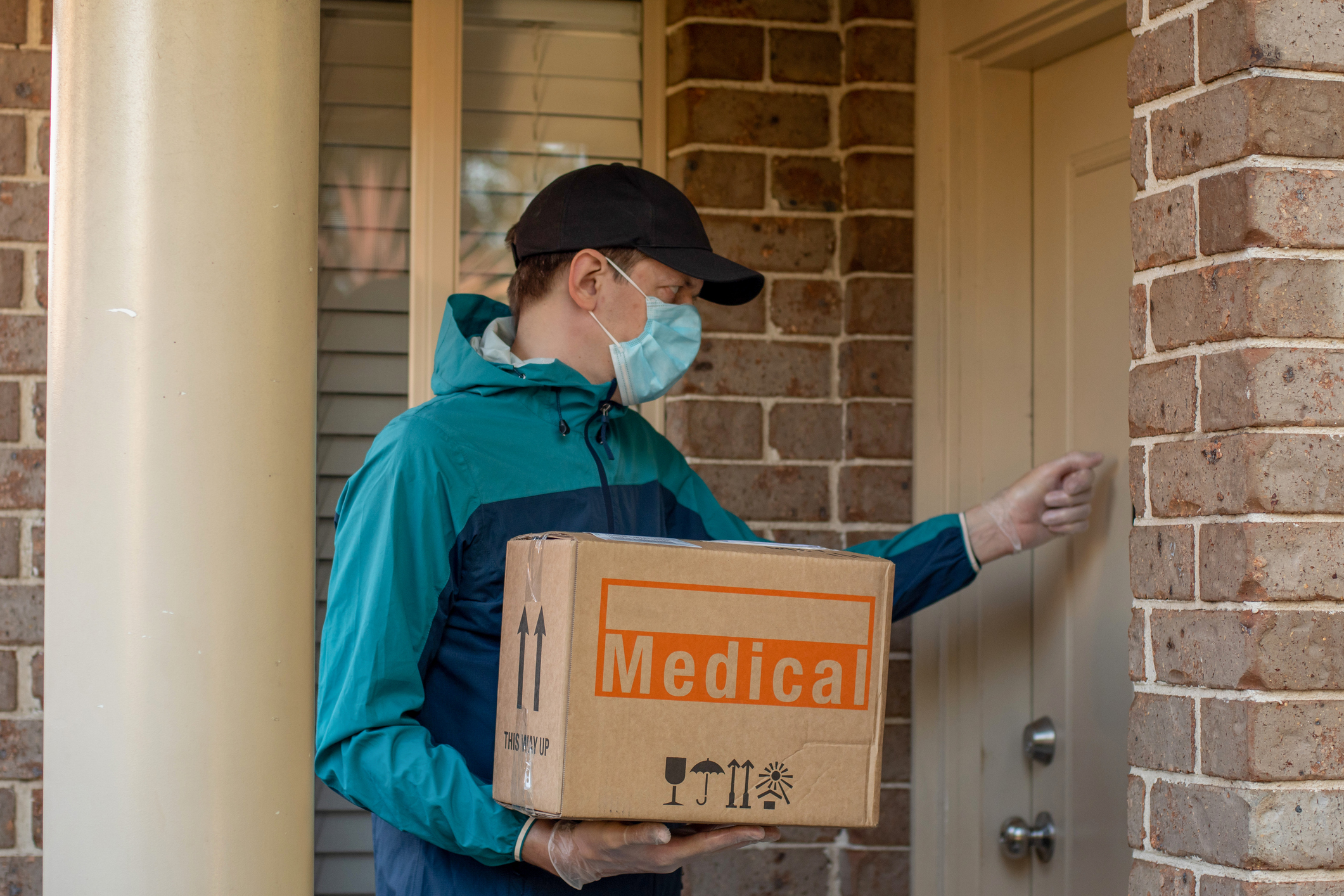 Contactless delivery during COVID-19 pandemic lockdown concept. Courier wearing mask and gloves holds a parcel with medical equipment and knocks on the door.